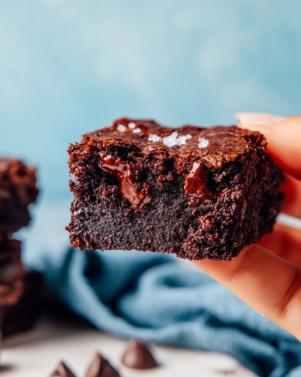 Several square brownies with a rich dark brown color and a slightly shiny, textured top layer are placed on a white marbled surface. Some brownies have smooth, round chocolate chips embedded on their top, adding a bit of contrast in gloss. The brownies are arranged closely, forming a rough grid, with a few pieces partially out of frame. On the top right, there is a round white bowl filled with many chocolate chips showing their white undersides, casting soft shadows inside. Scattered chocolate chips lie near the bowl and brownies. At the bottom left, a small area of blue cloth adds a soft touch to the scene. Photo taken with an iphone --ar 4:5 --v 7