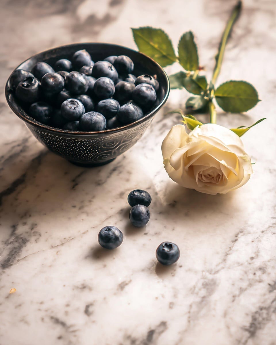A dark patterned bowl filled with many fresh blueberries and some green leaves is placed on a white marbled surface. Next to the bowl, a pale cream-colored rose with green sepals lays with its stem stretching out. Several loose blueberries are scattered in front of the bowl on the marbled surface. The image is softly lit, highlighting the textures of the blueberries, the petals of the rose, and the marble beneath. photo taken with an iphone --ar 4:5 --v 7