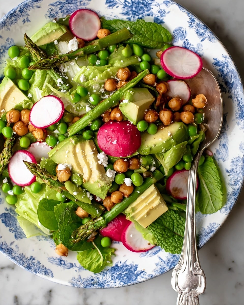 A salad on a white plate with blue floral patterns, with layers including bright green lettuce leaves as the base, scattered green peas and asparagus spears, round slices of pink and white radish, chunks of light green avocado, golden brown chickpeas, small red radishes, and white crumbled cheese sprinkled throughout. Fresh green mint and basil leaves are placed on top, adding texture and color contrast. Two silver spoons rest on the right side of the plate, and a small glass jar with green dressing and a spoon is placed nearby on a white marbled surface. photo taken with an iphone --ar 4:5 --v 7