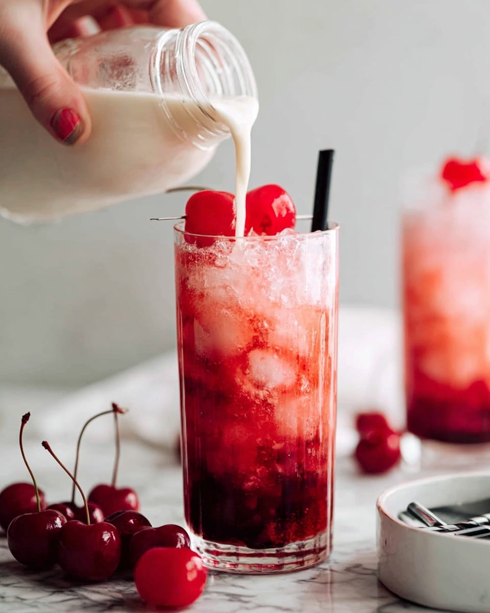 A tall clear glass filled with crushed ice holds a red drink with darker red layers swirling through it. On top of the ice are three bright red cherries with stems. A woman's hand is pouring creamy white liquid from a small glass jar into the glass, creating a soft white foam at the top. A black straw sticks out from the left side of the glass. Several bright red cherries with stems lie scattered on a white marbled surface around the glass, and parts of a white bowl and black straws are visible on the right side. In the background, a similar drink sits slightly out of focus. photo taken with an iphone --ar 4:5 --v 7