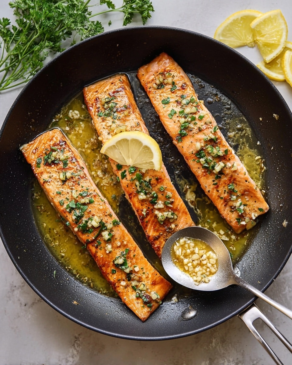 The image shows three thick salmon fillets cooked in a pan with a golden brown crust on top, sprinkled with small green herb pieces and tiny bits of garlic. Each fillet is topped with a small wedge of bright yellow lemon. The salmon has a shiny, slightly oily surface with a rich orange-pink color underneath the crust. The pan is dark and speckled, with a spoon filled with a light yellow sauce or oil resting on the side next to a couple of lemon slices. The background is a white marbled texture. photo taken with an iphone --ar 4:5 --v 7