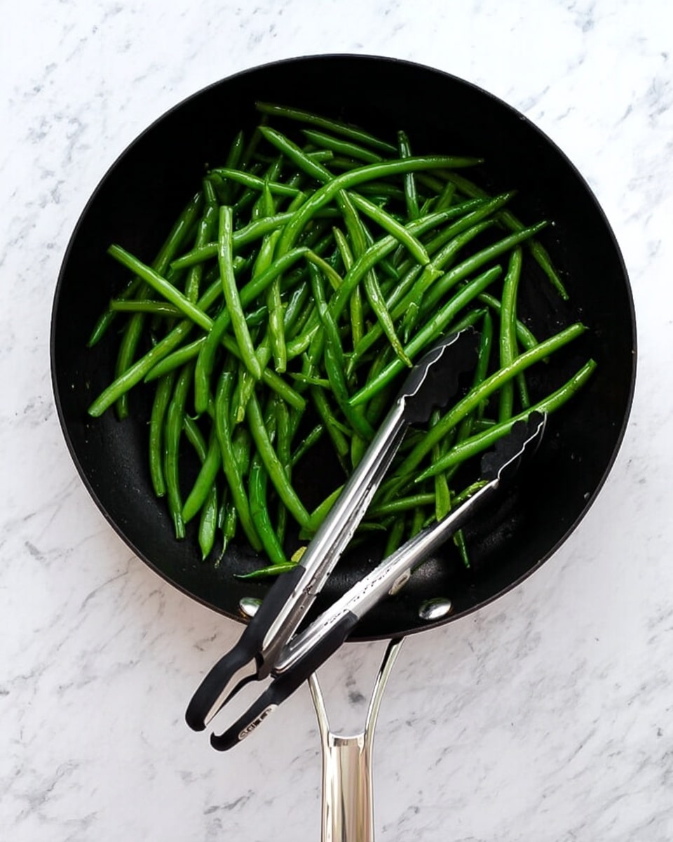 A close-up view of a white bowl filled with bright green beans, each bean smooth and glossy with small white pieces of garlic sprinkled evenly on top. The green beans are long and slender, layered loosely inside the bowl, with some beans overlapping each other. A silver fork rests on the edge of the bowl, its tines partially visible, giving a sense of scale and readiness to eat. The bowl sits on a white marbled surface, creating a clean and fresh setting. Photo taken with an iphone --ar 4:5 --v 7