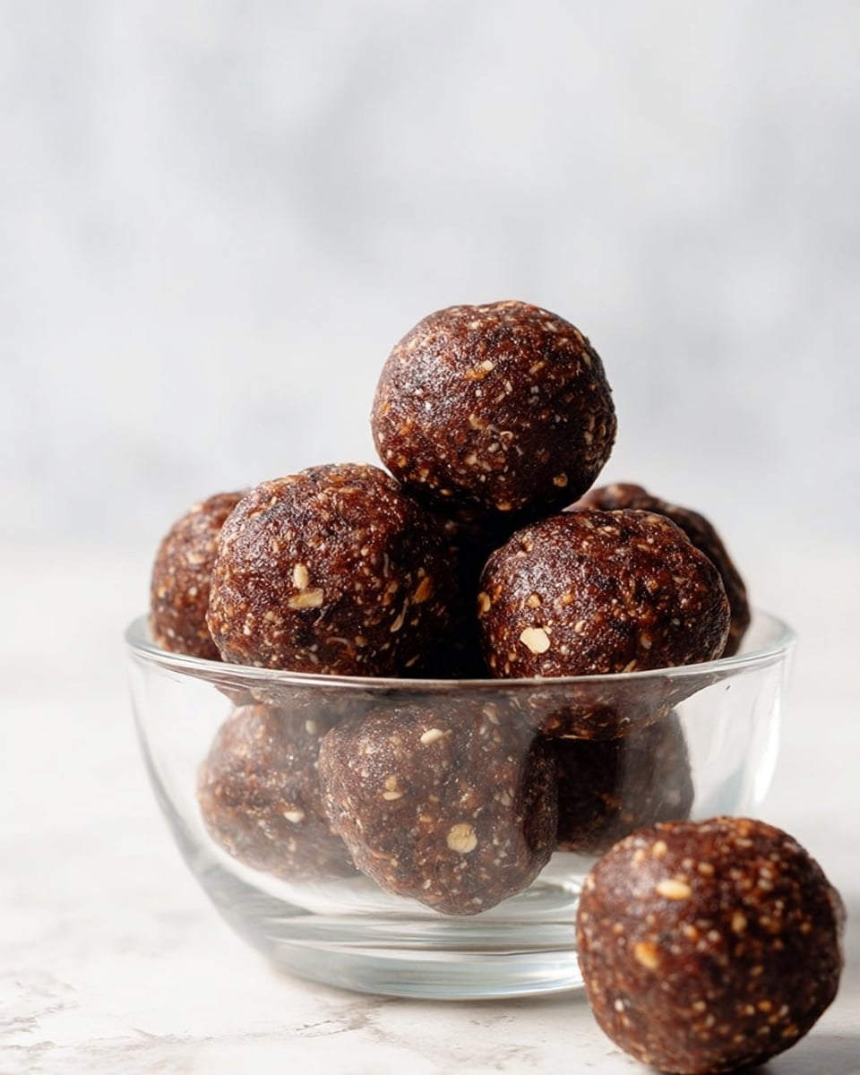 The image shows a clear glass bowl filled with round dark brown energy balls that have small visible bits of nuts or oats. The energy balls have a rough texture and appear firm. There are six balls inside the bowl, with some stacked on top of each other, and one ball placed just outside the bowl on a white marbled surface. The background is soft and blurred with the white marbled texture continuing in the background, giving a clean look. photo taken with an iphone --ar 4:5 --v 7