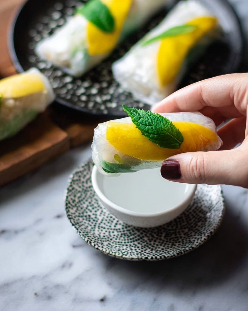 A woman's hand is holding a translucent spring roll filled with a bright yellow slice and a fresh green leaf near a small white bowl filled with a white liquid. The spring roll appears to have thin, clear rice paper wrapping, showcasing the yellow and green inside. In the background, there is a white patterned plate with more spring rolls topped with the same yellow slices and green leaves, all placed on a white marbled surface. Photo taken with an iphone --ar 4:5 --v 7