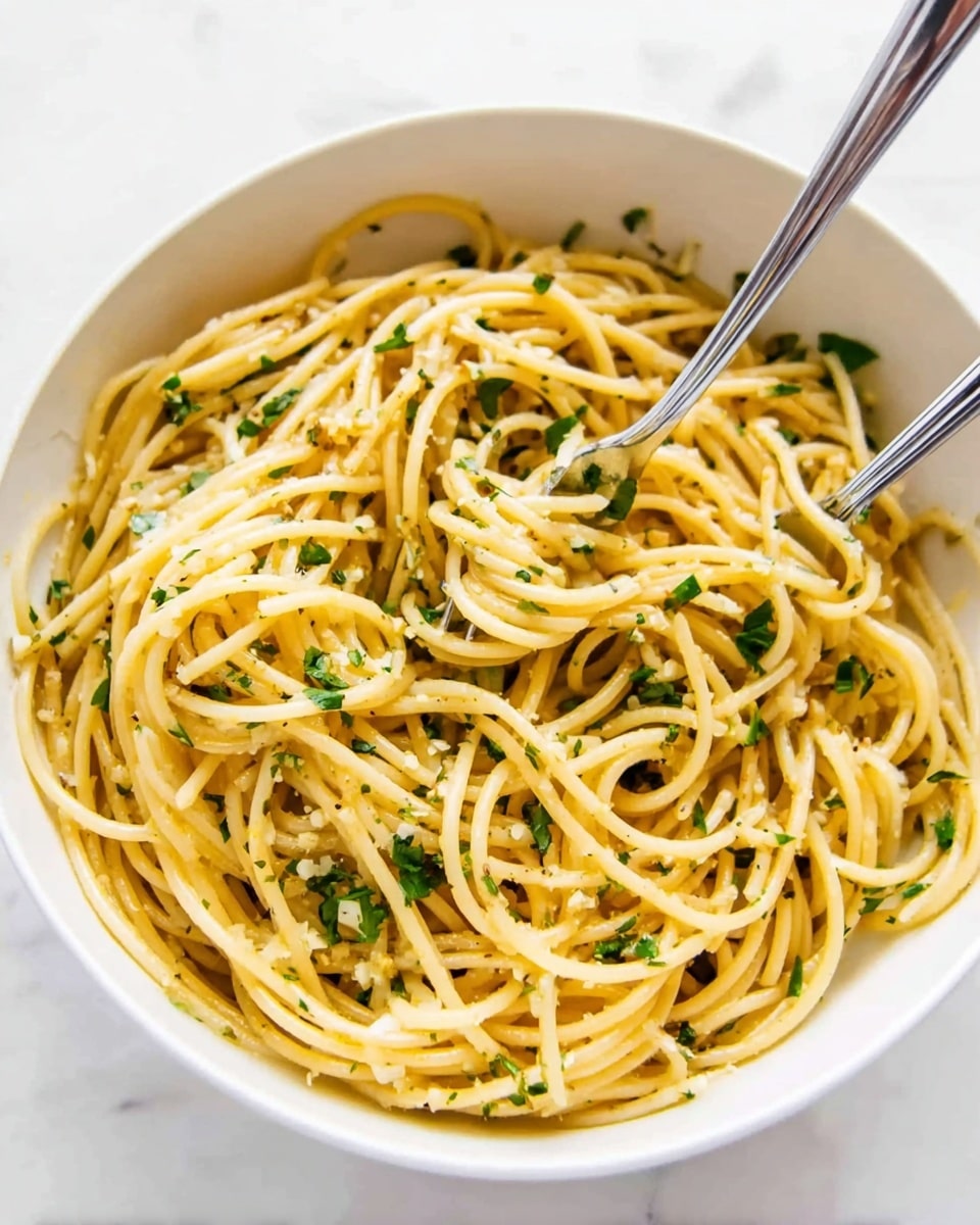 A large stainless steel pot filled with a single layer of cooked spaghetti noodles mixed with small pieces of chopped green herbs and thin slices of translucent cooked garlic. The pasta is light yellow, with the green herbs scattered evenly throughout, resting against the shiny inner surface of the pot. The pot is placed on a white marbled surface, and no other objects are visible in the image. Photo taken with an iphone --ar 4:5 --v 7