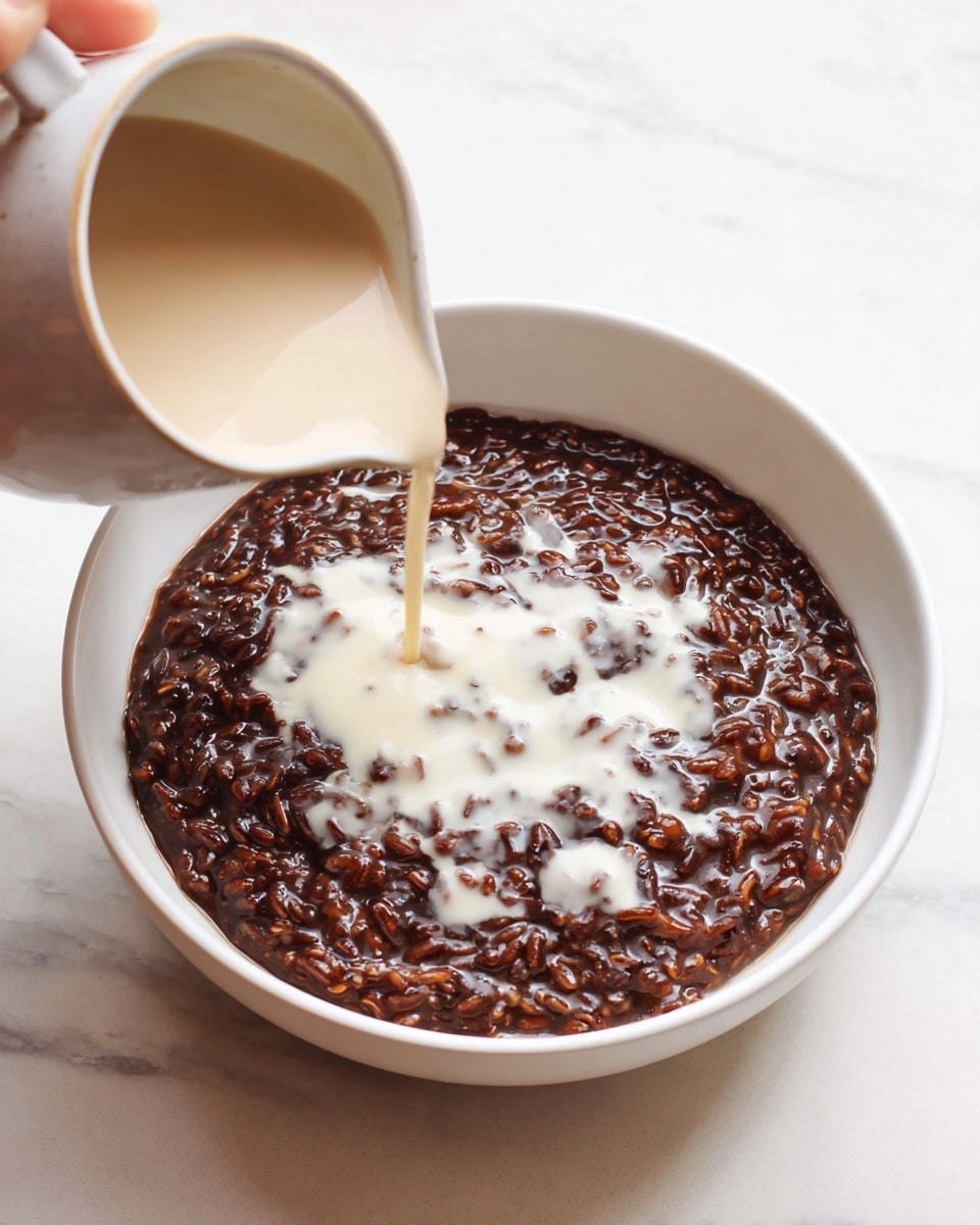 A white bowl holds a dark brown, thick rice pudding with a shiny and slightly sticky texture, filled with visible grains of rice. From above, a light beige creamy liquid is being poured onto the center of the pudding from a ceramic pitcher held by a woman's hand, creating a contrast between the dark base and the pale cream forming irregular shapes on top. The bowl sits on a white marbled surface, enhancing the clean and simple presentation. photo taken with an iphone --ar 4:5 --v 7