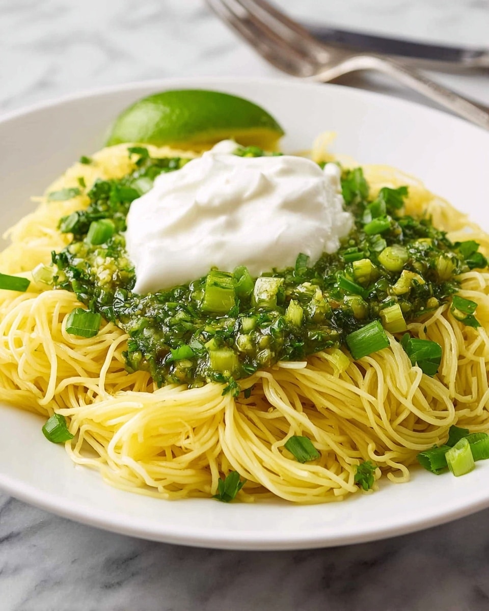 A white bowl holds a nest of thin, pale yellow noodles mixed with small pieces of green herbs and spring onions. In the center, there is a layer of chunky, bright green sauce topped with a dollop of smooth, white cream. A green lime wedge rests on the right side of the noodles, next to a silver spoon, all set on a white marbled surface with a bright green cloth nearby. photo taken with an iphone --ar 4:5 --v 7