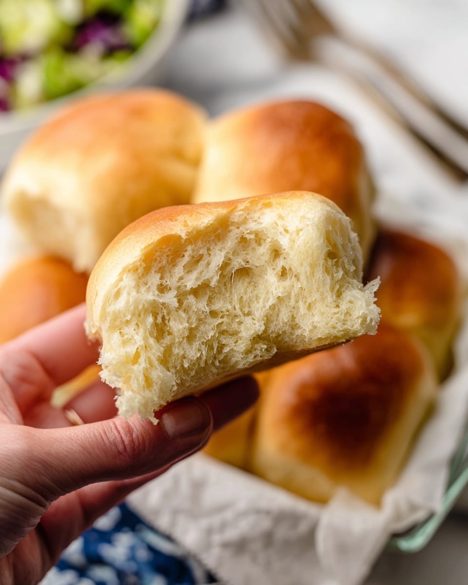 A close-up image shows several golden brown, soft dinner rolls with a light texture. The rolls are stacked in a white basket lined with a white and black striped cloth, with one roll torn open revealing its fluffy inside. In the blurry background, there is a white marbled surface and a hint of a colorful salad in a white bowl. The warm colors and soft texture make the rolls look fresh and inviting. photo taken with an iphone --ar 4:5 --v 7