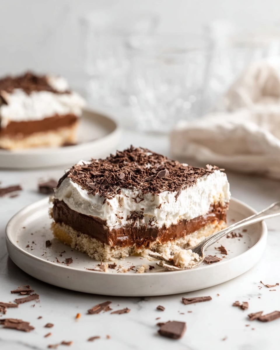 A square piece of layered dessert sits on a white plate with a silver fork holding a small bite next to it. The dessert has three main layers: a light beige crumbly base, a thick middle layer of smooth, rich chocolate, and a top layer of fluffy white cream. The dessert is covered with dark chocolate shavings that add texture and depth. Scattered chocolate pieces and crumbs are around the plate, placed on a white marbled surface. In the background, a second plate with another piece of dessert and blurred clear glasses are visible. photo taken with an iphone --ar 4:5 --v 7