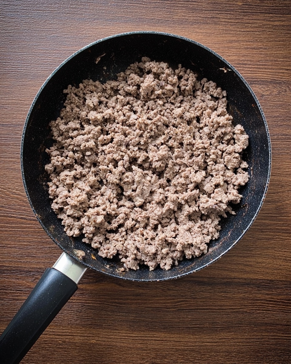 A black frying pan with a long handle is shown from above, filled with evenly cooked minced meat spread in one thick layer covering the whole pan. The texture of the meat is crumbly and brownish-gray, with small bits clumped together, giving a rough and granular look. The pan sits on a brown wooden surface with a simple pattern. photo taken with an iphone --ar 4:5 --v 7