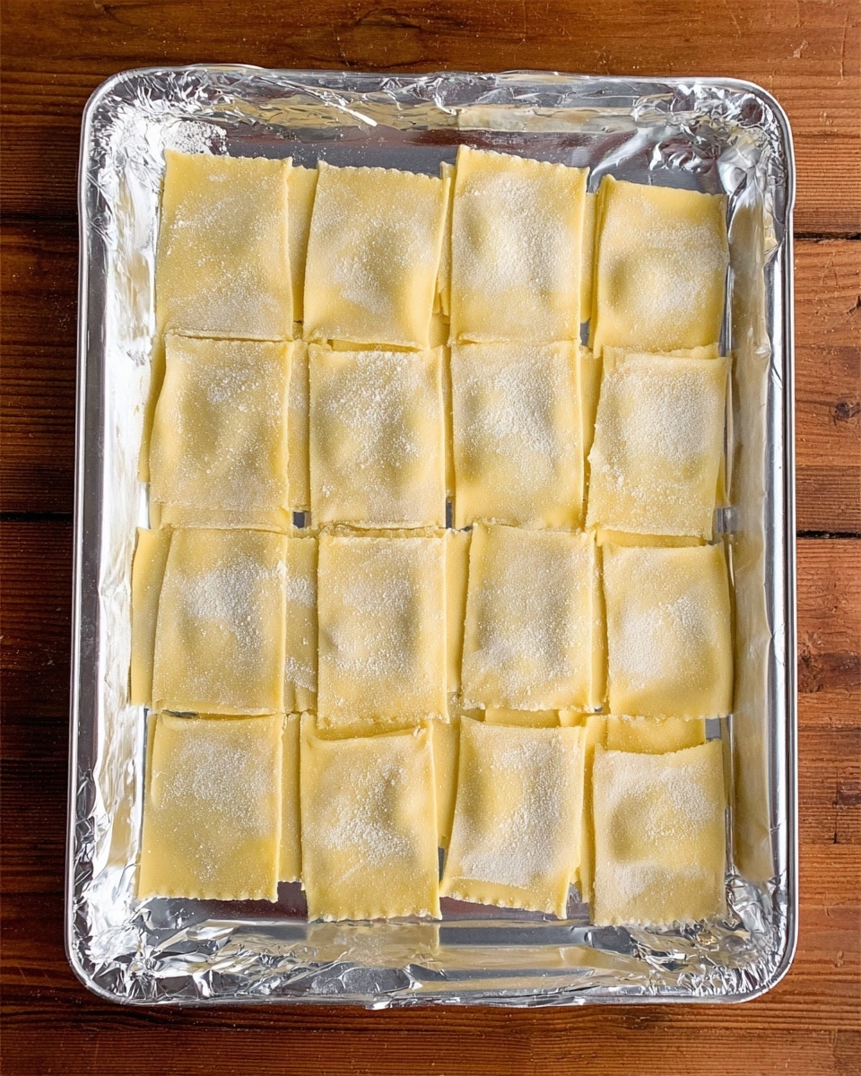 A metal tray covered with shiny aluminum foil is placed on a wooden surface. On the tray, there are 27 rectangular sheets of uncooked pasta dough, arranged in three neat rows. The pasta sheets have a pale yellow color and a smooth texture, with a light dusting of white flour on top. The edges of some sheets are slightly uneven, showing they were manually prepared. The lighting is bright and natural, highlighting the soft texture of the dough photo taken with an iphone --ar 4:5 --v 7