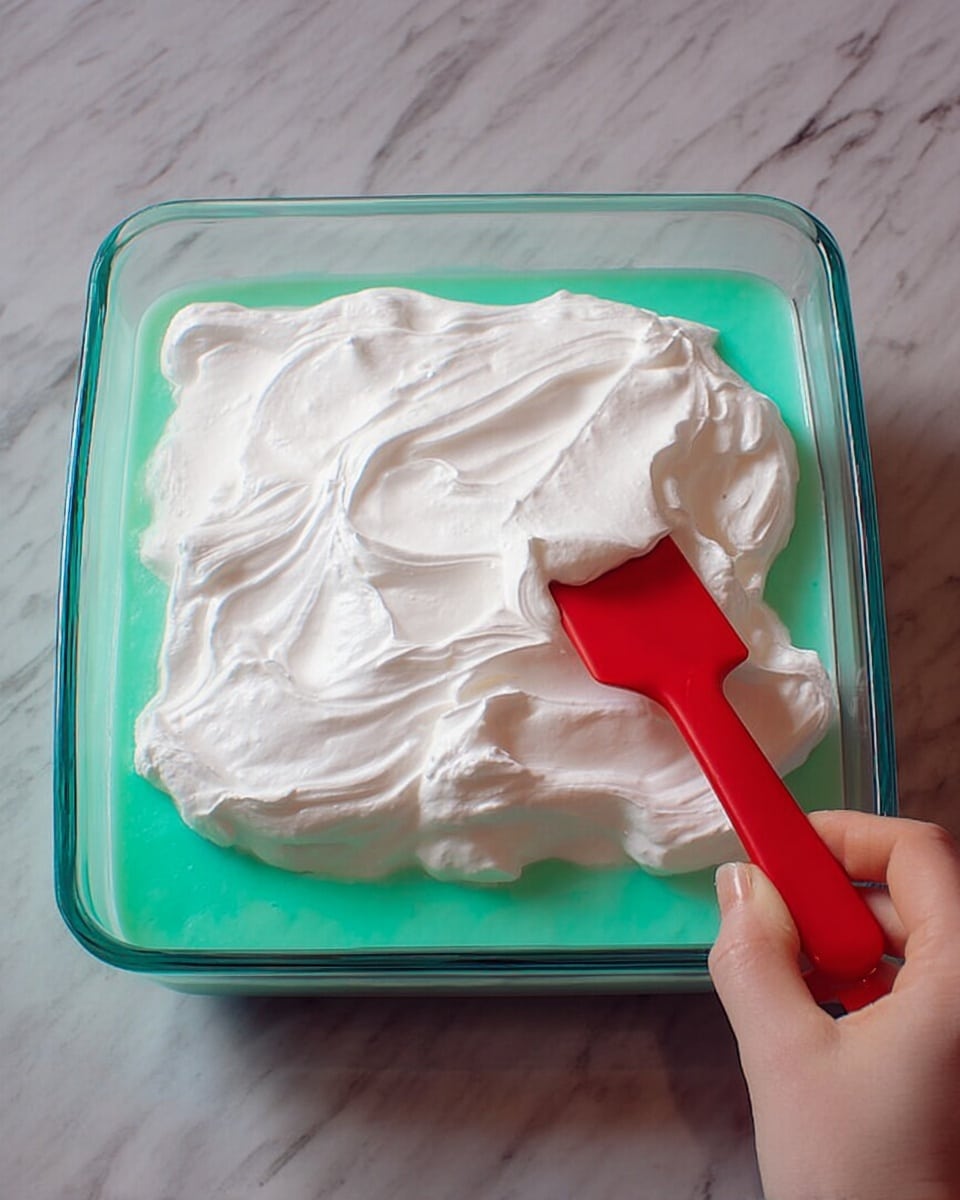 The image shows a square glass dish filled with two distinct layers of food. The bottom layer is a smooth, light turquoise green mixture that fills about half the dish. On top of this, there is a thick white layer of fluffy whipped topping covering the surface unevenly with gentle folds and swirls. A woman's hand is holding a red spatula that is spreading the white topping over the green layer, creating a contrast between the colors and textures. The dish is placed on a surface with a white marbled texture. photo taken with an iphone --ar 4:5 --v 7