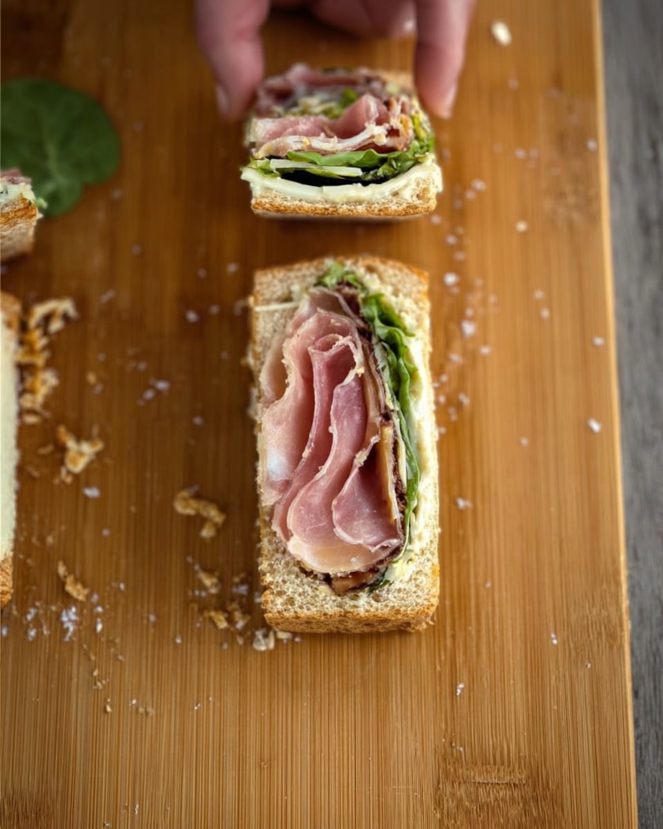 A close-up view of a wooden board with a sandwich roll that has a rectangular shape on the right side and two small round pieces sliced on the left side. The sandwich layers include a light brown toast base, a creamy spread layer, green leafy vegetables, a thin layer of white cheese, and a folded slice of pink deli meat on top. The small round slices reveal all these layers with the dark green and light green leaves visible, and the sandwich is sprinkled with crumbs scattered across the wooden board. A woman's hand can be seen holding one of the round sandwich slices on the left side. photo taken with an iphone --ar 4:5 --v 7