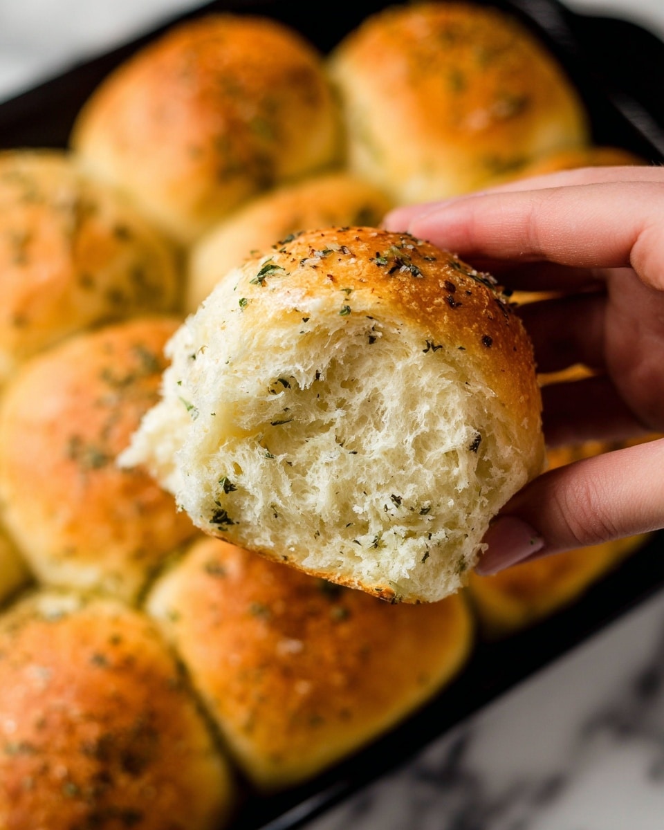 The image shows a close-up of a cast iron pan filled with a single layer of round, golden brown dinner rolls. Each roll is topped with coarse sea salt flakes and small green herb pieces, creating a textured surface. The rolls are soft and tightly packed, with light cracks and a slightly shiny crust from baking. The pan sits on a white marbled surface, and a green cloth is partially visible in the background. photo taken with an iphone --ar 4:5 --v 7