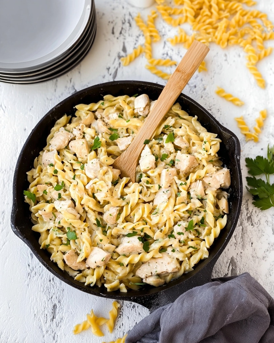 The image shows a close-up of a black cast-iron skillet filled with a pasta dish. The pasta is short and twisted with a pale yellow color, mixed with small chunks of cooked white meat with a tender texture. Green parsley leaves are scattered on top, adding a splash of color. A wooden spoon with a light wood tone is placed inside the skillet, partly buried in the food, positioned upright. Around the skillet, there are some uncooked pasta pieces and a gray cloth draped casually over the edge of the skillet handle. The background surface is a white marbled texture, with a few stacked light gray plates visible in the upper left corner. Photo taken with an iphone --ar 4:5 --v 7