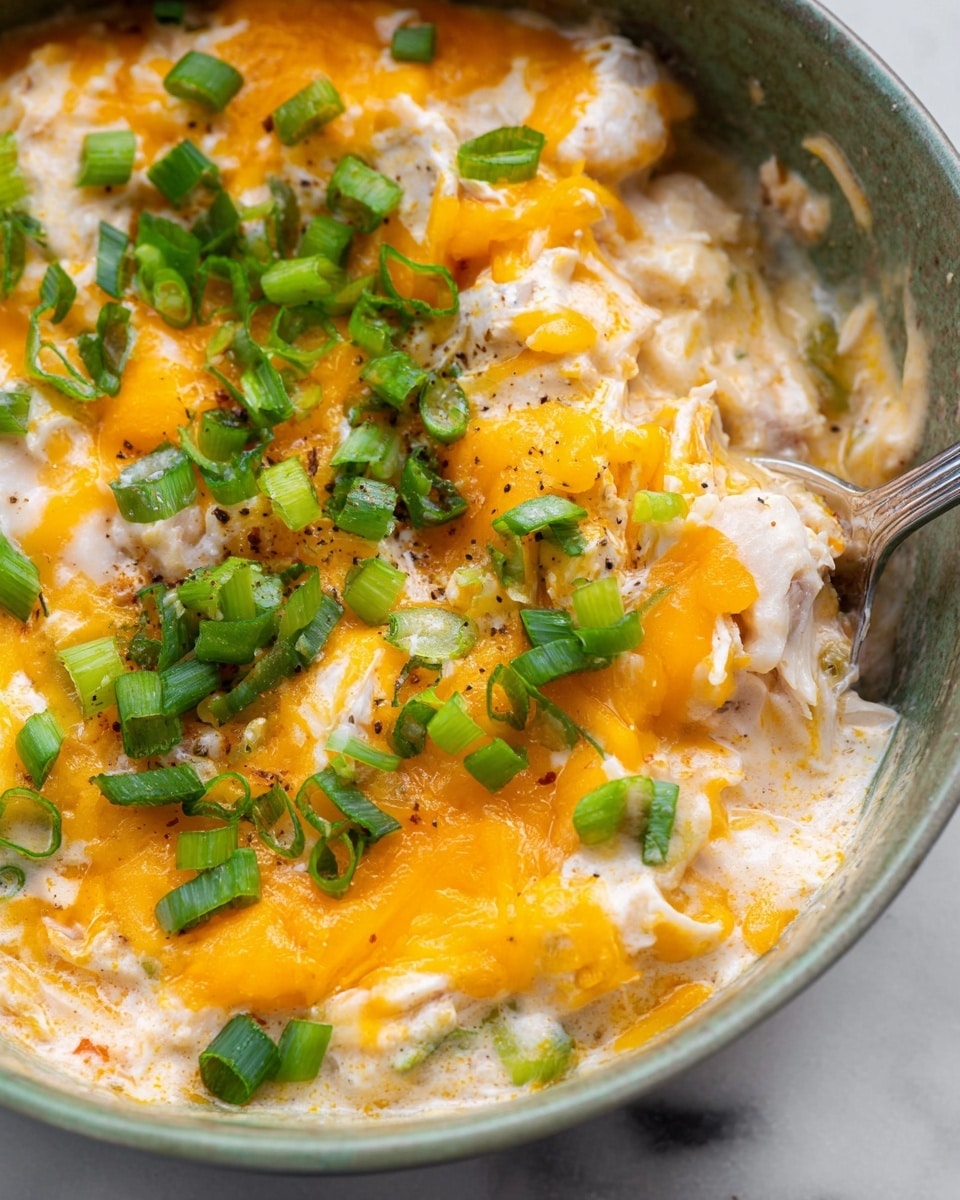 This is a close-up view of a creamy dish with multiple layers in a white bowl. The base layer is a light orange creamy sauce mixed with white shredded pieces. On top of this, there are bright yellow melted cheese layers spread around unevenly, blending slightly with the creamy sauce. Scattered over the top, there are fresh chopped green onions adding a fresh green color contrast. A silver spoon is partially visible, scooping some of the creamy mixture. The background is a white marbled texture. photo taken with an iphone --ar 4:5 --v 7