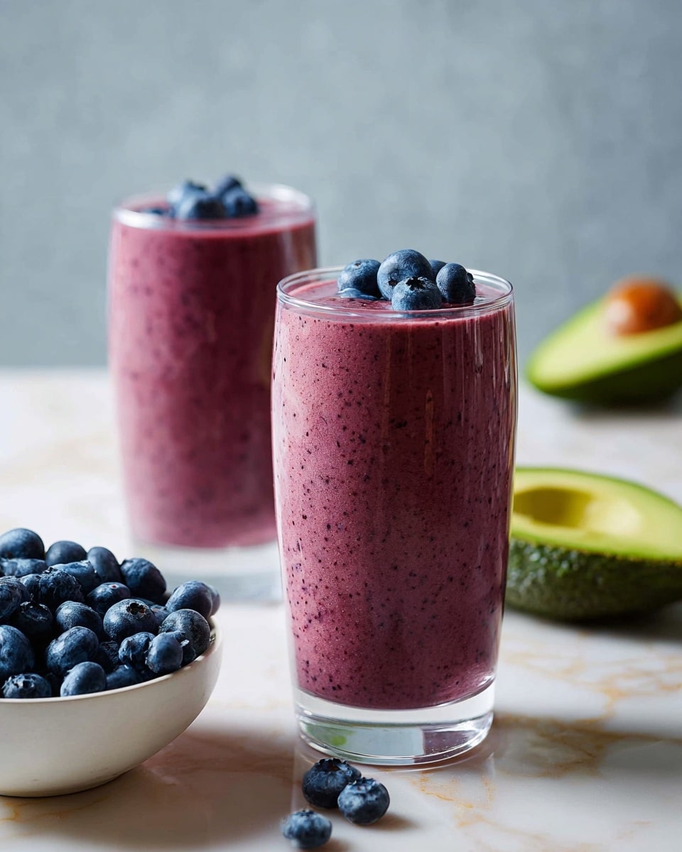 Two tall clear glasses are filled with smooth, thick purple smoothie with tiny dark specks visible inside. Each glass is topped with three fresh blueberries sitting on the smoothie surface. Behind the glass in front, there is a white bowl with many blueberries. The smooth background and surface have a white marbled texture, and part of a halved avocado with seed is visible on the lower left side. Photo taken with an iphone --ar 4:5 --v 7
