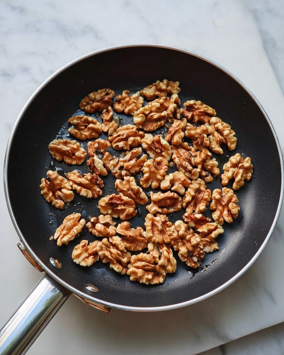 The image shows a black frying pan filled with many walnut halves that are lightly toasted with some darker spots, giving them a warm brown color and a slightly rough texture. The pan has a metal handle and is placed on a white marbled surface, which provides a clean and bright background that contrasts with the darker pan and nuts. photo taken with an iphone --ar 4:5 --v 7