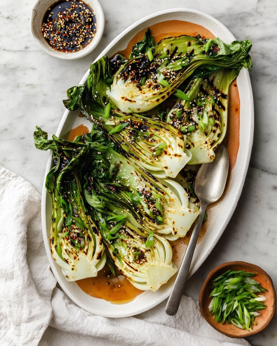 The image shows four grilled bok choy halves arranged on a large oval white plate with a warm orange tint. Each bok choy has dark green, slightly charred leafy edges and pale green to white inner stalks with visible layers. They are sprinkled with black sesame seeds, small red chili flakes, and chopped bright green scallions, adding texture and color contrast. To the side of the plate, there is a silver serving spoon partially resting on the bok choy. In the background, a small white bowl holds a dark sauce with minced garlic, and a small round bowl contains green scallions and black sesame seeds. The whole setting is on a white marbled surface with a white cloth nearby. photo taken with an iphone --ar 4:5 --v 7