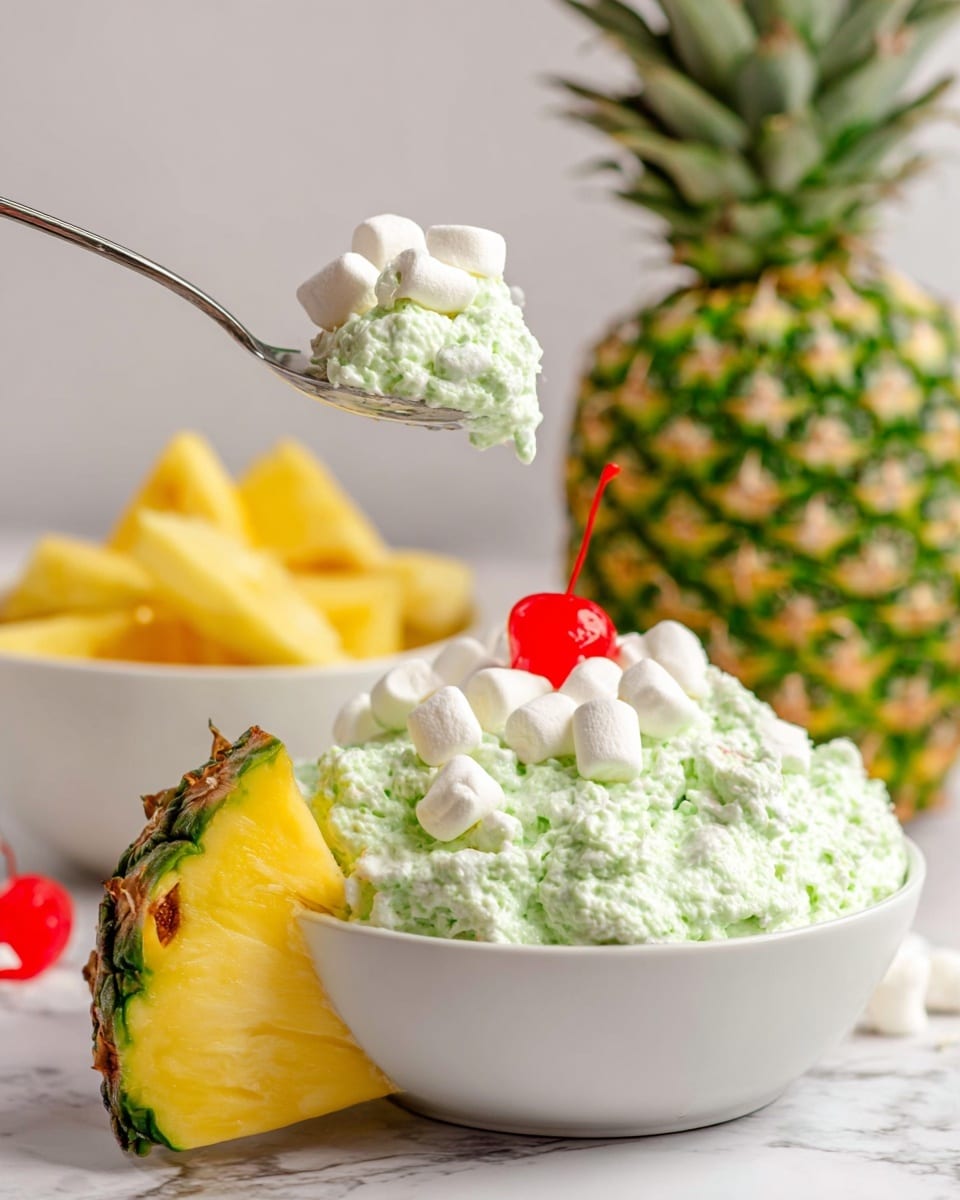 A white bowl filled with a light green fluffy salad that has a creamy texture with small visible bits inside. On top of the salad, there are five white marshmallows scattered around a bright red cherry with a long stem in the center. A thick triangular slice of pineapple with a rough green and yellow rind is placed standing inside the salad next to a silver spoon handle sticking out of the bowl. In the background, there is a whole pineapple with a rough green and brown skin and another white bowl with more of the same green salad. The setting is on a white marbled surface photo taken with an iphone --ar 4:5 --v 7