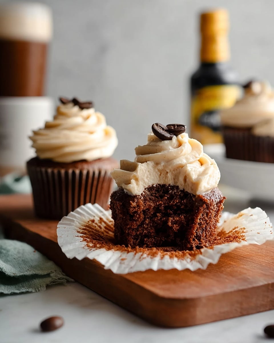 The image shows two chocolate cupcakes on a small wooden board lined with white parchment paper. Each cupcake has a dark brown base with a swirl of light beige frosting on top, finished with a single dark coffee bean placed in the center of the frosting. In the background, there are more cupcakes on white surfaces, a bottle with a yellow label, a small white bowl with coffee beans, and a white vase holding dried plants, all set on a white marbled surface. Scattered coffee beans and a spoon with a wooden handle also appear around the board. photo taken with an iphone --ar 4:5 --v 7