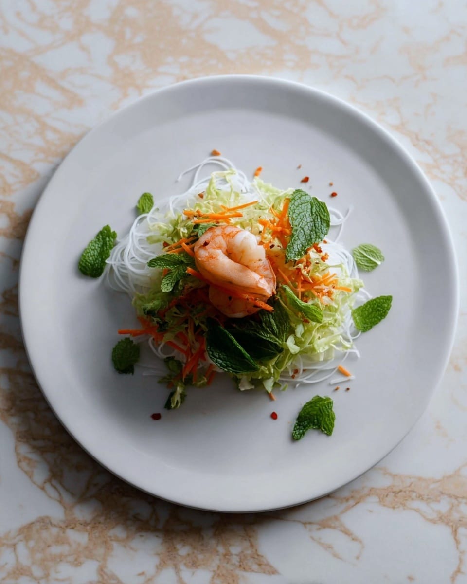A small serving of food is arranged neatly in the center of a large white plate. The bottom layer is thin, white rice noodles curled in a loose pile. On top of the noodles is a layer of shredded light green cabbage and bright green mint leaves scattered around. A single cooked shrimp, pale pink with white stripes, sits in the middle. Finely shredded orange carrot is sprinkled over the shrimp and greens. The plate sits on a white marbled surface with subtle beige patches. photo taken with an iphone --ar 4:5 --v 7