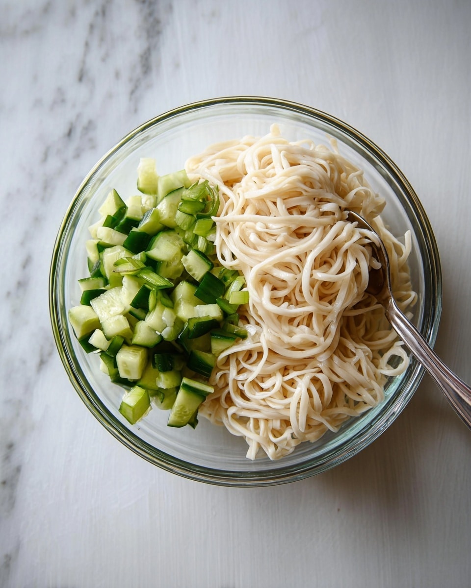 A clear glass bowl on a white marbled surface contains two main layers: on the left side there are small, bright green cucumber pieces cut into cubes with smooth edges, and on the right side there is a large pile of thick, pale off-white noodles that look soft and slightly twisted. A silver spoon rests inside the bowl on the noodle side, angled toward the bottom right. The overall look is clean and fresh with a simple arrangement photo taken with an iphone --ar 4:5 --v 7