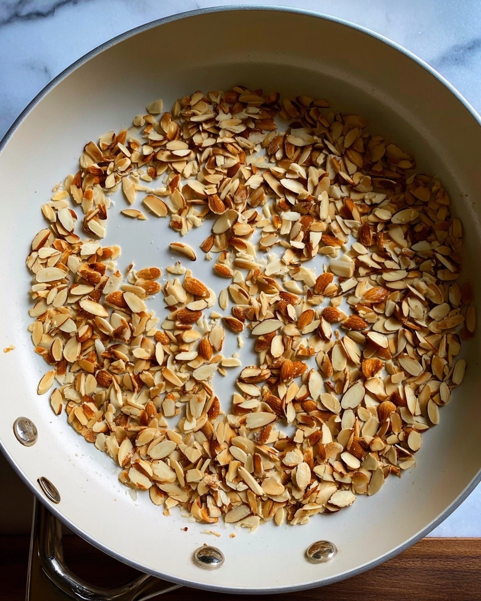 A large white pan filled with many small, toasted almond slices scattered unevenly across the bottom. The almond slices are light brown with darker toasted spots, giving a mix of golden and amber shades. The pan itself has a smooth, clean surface with metal rivets on the edges visible. The background is a white marbled texture. photo taken with an iphone --ar 4:5 --v 7