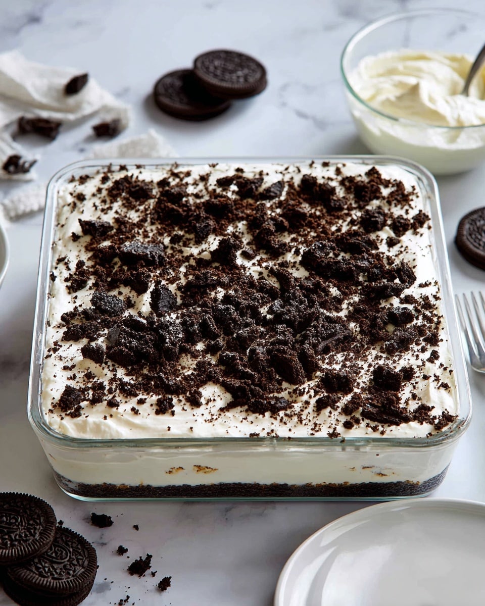 A square glass pan filled with a layered dessert. The bottom layer is not visible but likely solid, topped by a thick white creamy layer that covers the entire pan evenly. The top layer consists of dark brown crushed cookie pieces scattered all over, giving a rough texture and contrast to the smooth white cream. Around the pan, there are whole dark chocolate cookies placed on a white marbled surface, and a clear glass bowl with white cream beside it. A white plate is partially visible on the right. The background is clean with a soft white marbled texture. photo taken with an iphone --ar 4:5 --v 7