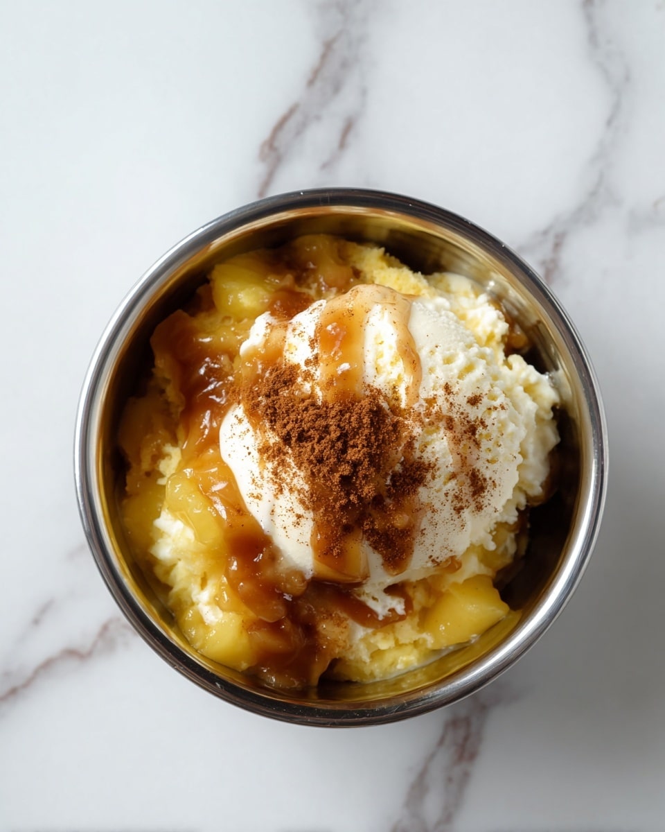 The dessert is served in a clear glass cup with three main layers visible from top view. The bottom layer is a creamy light yellow custard. On top is a layer of white whipped cream that looks soft and fluffy. The topmost layer has chunks of golden-brown cake pieces mixed with bright yellow pineapple slices and a shiny bright red cherry in the center. A drizzle of caramel sauce adds a glossy amber color over the cake and fruit. The glass cup is placed on a white marbled surface. Photo taken with an iphone --ar 4:5 --v 7