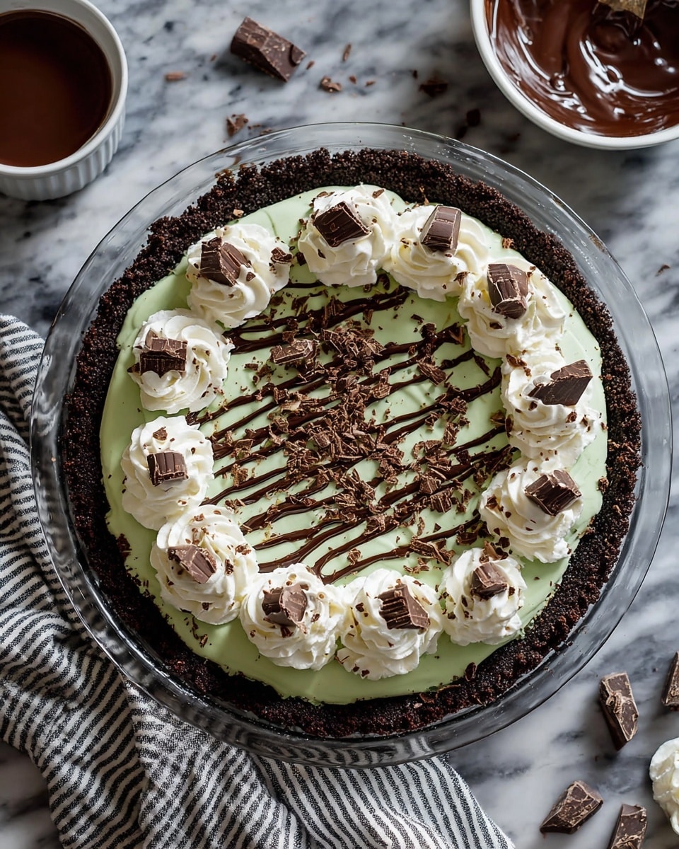 A pie in a clear glass pan has three main layers: the bottom is a dark chocolate crumb crust, the middle is a smooth pale green filling, and the top has white whipped cream dollops evenly spaced around the edge and one in the center. Each dollop is decorated with a small piece of chocolate. The green layer is drizzled with thin dark chocolate lines and sprinkled with broken chocolate pieces all over. The scene is set on a white marbled surface with a white bowl of melted chocolate nearby and some striped cloth under the pan. photo taken with an iphone --ar 4:5 --v 7