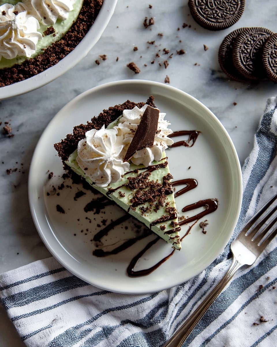 A close-up of a slice of layered dessert on a plain white plate stacked on another white plate, set on a white marbled surface. The dessert has a bottom layer of dark chocolate cookie crust, topped by a pale mint green creamy layer mixed with small dark cookie bits. Above this is a thick white creamy layer. The top is decorated with swirls of white whipped cream, with small broken dark cookie pieces and a striped chocolate mint candy stick standing upward on one side. Drizzles of dark chocolate syrup run down from the whipped cream. The background shows a blurred second plate with a similar dessert and a white container. Photo taken with an iphone --ar 4:5 --v 7