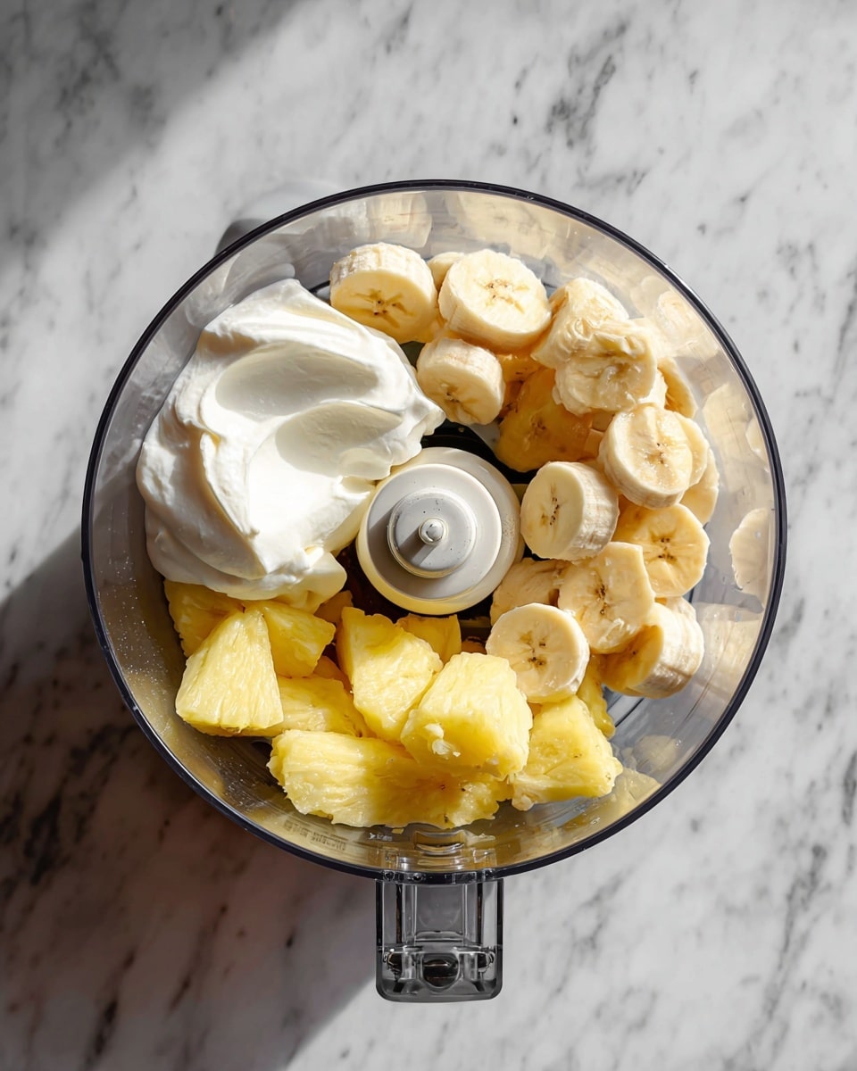 A clear food processor bowl sits on a white marbled surface, filled with three main layers: at the bottom are pale yellow frozen pineapple chunks with a bumpy texture, on top of them are round, smooth slices of light yellow banana, and on one side, there is a large dollop of white creamy coconut cream. The blade of the food processor is visible through the bowl, positioned centrally near the ingredients. photo taken with an iphone --ar 4:5 --v 7