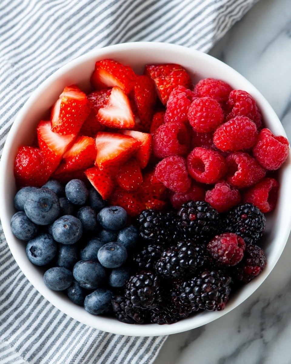 A white bowl filled with four clear sections of fresh fruit on a white marbled surface. The top left section is filled with bright red strawberry halves, showing their juicy texture and white inner flesh. The bottom left section holds small, round, deep blue blueberries with a smooth surface. The bottom right section contains vibrant red raspberries, each with a bumpy and hollow texture. The top right section has shiny dark blackberries with a textured, clustered appearance. The background shows a faintly visible white and grey striped cloth. photo taken with an iphone --ar 4:5 --v 7