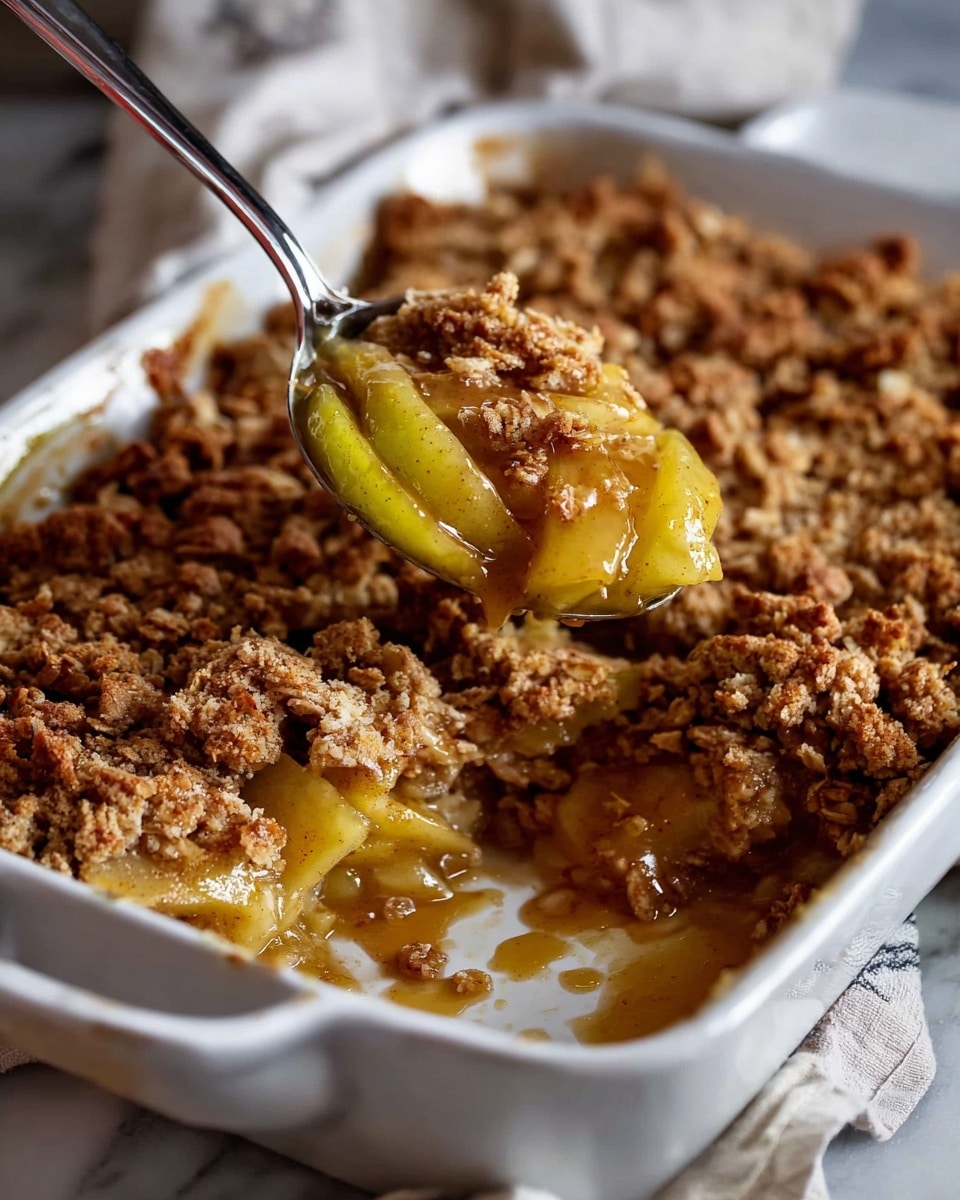 A rectangular white baking dish filled with a golden brown apple crumble that has a crumbly, textured top layer with visible oats and small chunks of baked apples peeking through. The dessert surface shows slight bubbling caramelized sauce around the edges. The dish is placed on a white cloth with red stripes, resting on a wooden board with a green apple and two cinnamon sticks nearby, all set against a dark white marbled surface. photo taken with an iphone --ar 4:5 --v 7