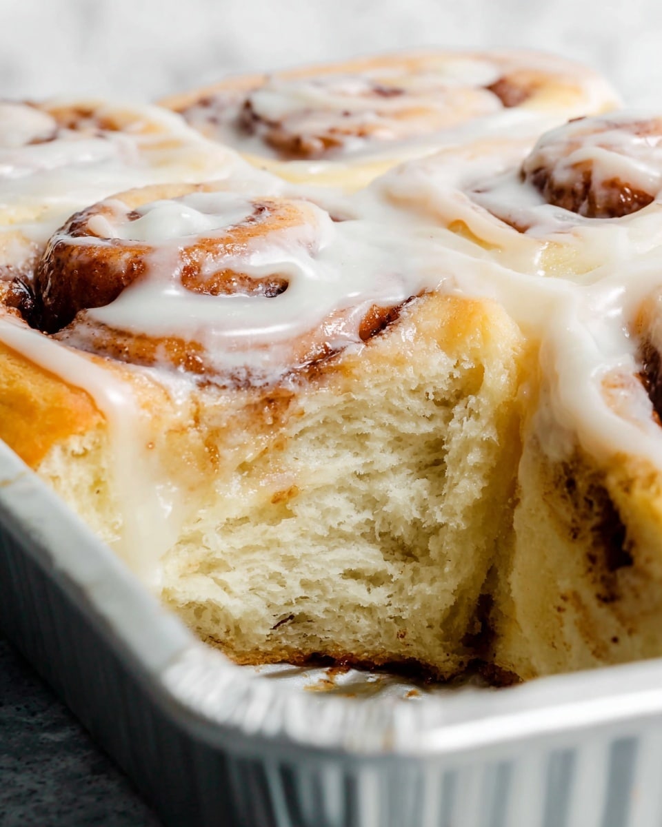 A close-up of a cinnamon roll being held by a person's fingers, showing the roll's spiral shape with three visible layers of soft, light brown dough and cinnamon filling. The top layer is covered with a thick, creamy white icing that looks smooth and slightly glossy, melting down the sides. The background shows more cinnamon rolls blurred on a white marbled surface. Photo taken with an iphone --ar 4:5 --v 7