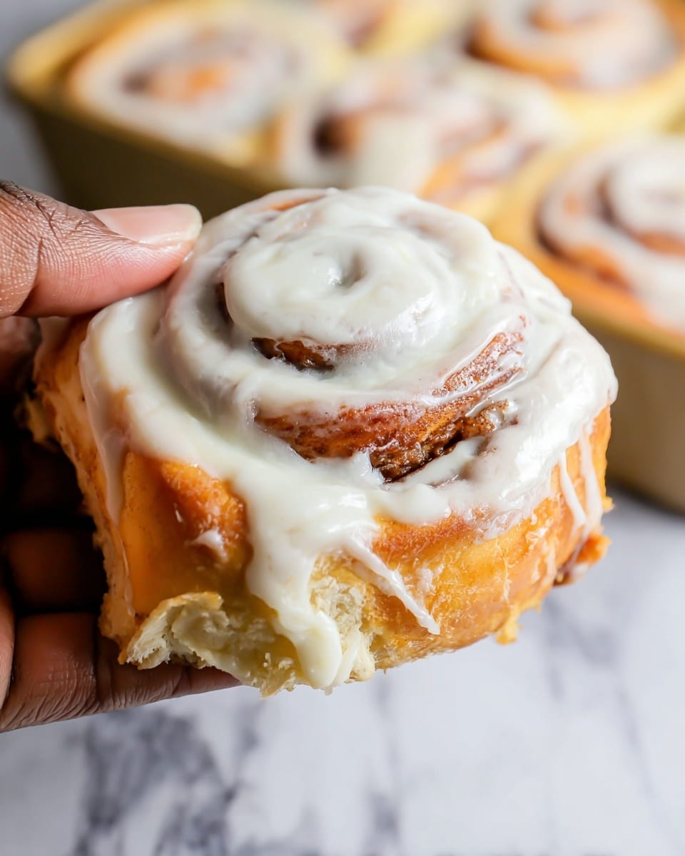 A close-up view of soft cinnamon rolls in a white tray lined with parchment paper, showing several swirled layers of light golden-brown dough and darker cinnamon filling. The rolls are thickly topped with a smooth, creamy white icing spread evenly across the swirls and pooling slightly at the edges. The tray sits on a white marbled surface, adding a clean and bright look to the image. Photo taken with an iphone --ar 4:5 --v 7