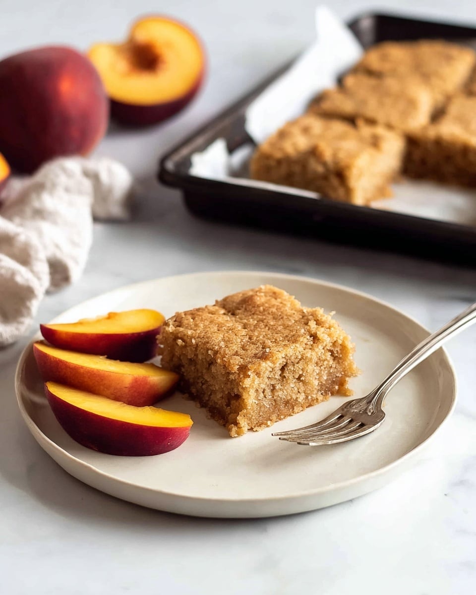 A single square piece of light brown cake with a rough, crumbly texture sits in the center of a white plate, accompanied by three thin slices of dark red and orange peach on the left side. To the right of the cake piece, a silver fork rests gently on the plate’s edge. Behind the plate, there is a whole peach and peach half revealing its yellow-orange inside. In the blurred background, a larger tray of similar cake squares is visible on white paper. The whole scene is set on a white marbled surface. Photo taken with an iphone --ar 4:5 --v 7
