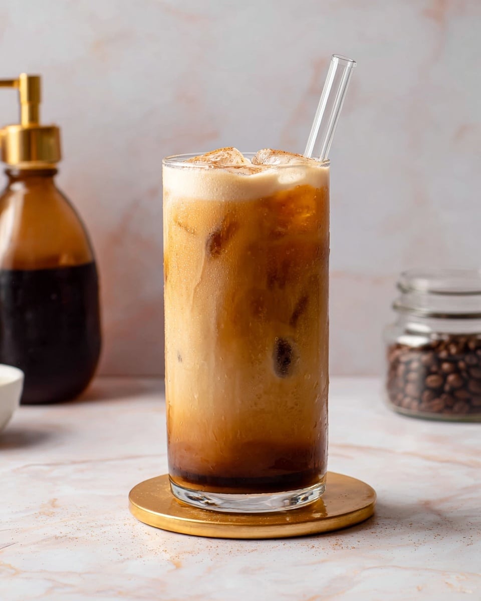 A tall clear glass filled with iced coffee showing three main layers: a darker coffee layer at the bottom, a creamy light brown layer in the middle with visible ice cubes, and a frothy light foam on top sprinkled with cinnamon. A clear glass straw stands upright from the foam. The glass sits on a round gold coaster placed on a white marbled surface. In the blurred background, there is a glass bottle with dark liquid and a golden pour spout on the left and a small white glass jar holding coffee beans on the right. Photo taken with an iphone --ar 4:5 --v 7