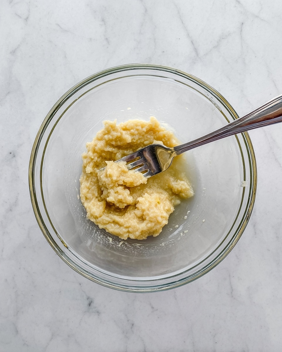 A clear glass bowl sits on a white marbled surface, containing a small portion of mashed banana that is light yellow with a slightly chunky texture. Inside the bowl, a silver fork rests on top of the mashed bananas, positioned horizontally across the mixture. The image is clear and simple, focusing on the single-layer of mashed banana in the bowl. photo taken with an iphone --ar 4:5 --v 7