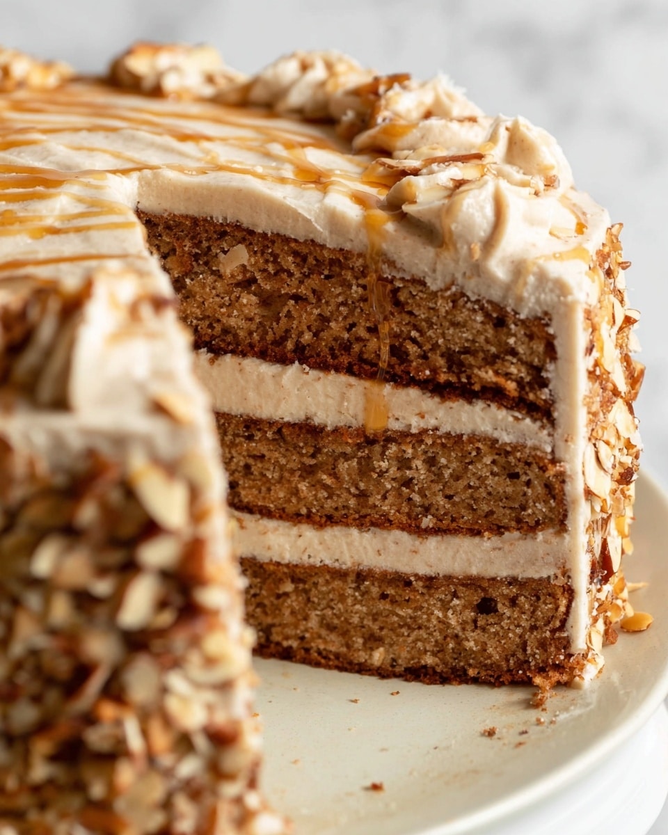 A round cake with two visible layers is covered in a light tan creamy frosting that has a smooth texture and a slightly rustic finish showing a bit of the cake through it. The top of the cake features a spiral pattern made with a light caramel or honey-colored glaze swirled evenly from center to edge. Around the bottom base of the cake, there is a thick ring of chopped nuts with a mix of beige and brown pieces. Additional nut pieces are scattered lightly on the very top edge, adding texture and detail. The cake sits on a white plate placed on a white marbled surface, and the background is softly blurred with some neutral tones of white and beige. photo taken with an iphone --ar 4:5 --v 7