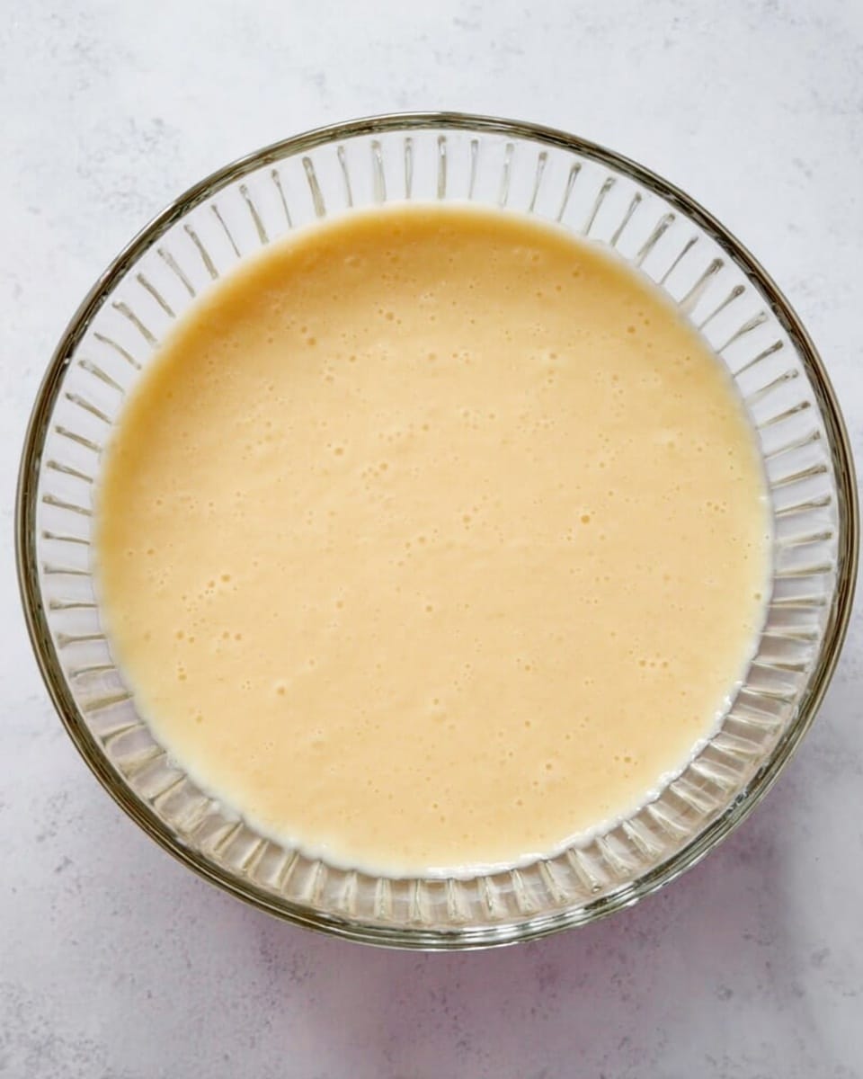 The image shows a clear glass bowl with a smooth, light yellow batter inside. The batter has a creamy texture with small bubbles and a slightly uneven surface. The bowl is placed on a white marbled surface, giving a clean and bright background that contrasts with the pale batter. The bowl’s ridged sides create soft shadows and add detail around the batter. photo taken with an iphone --ar 4:5 --v 7
