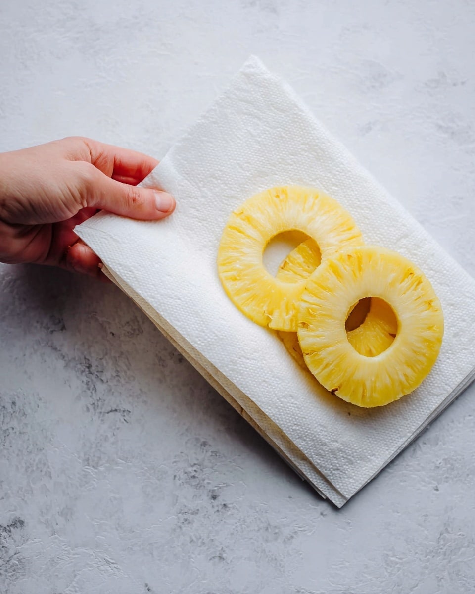 A woman's hand is lifting a corner of a stack of white paper towels that hold two yellow pineapple rings with a hole in the middle, showing another layer of pineapple rings under the paper towels. The stack rests on a white marbled surface. photo taken with an iphone --ar 4:5 --v 7