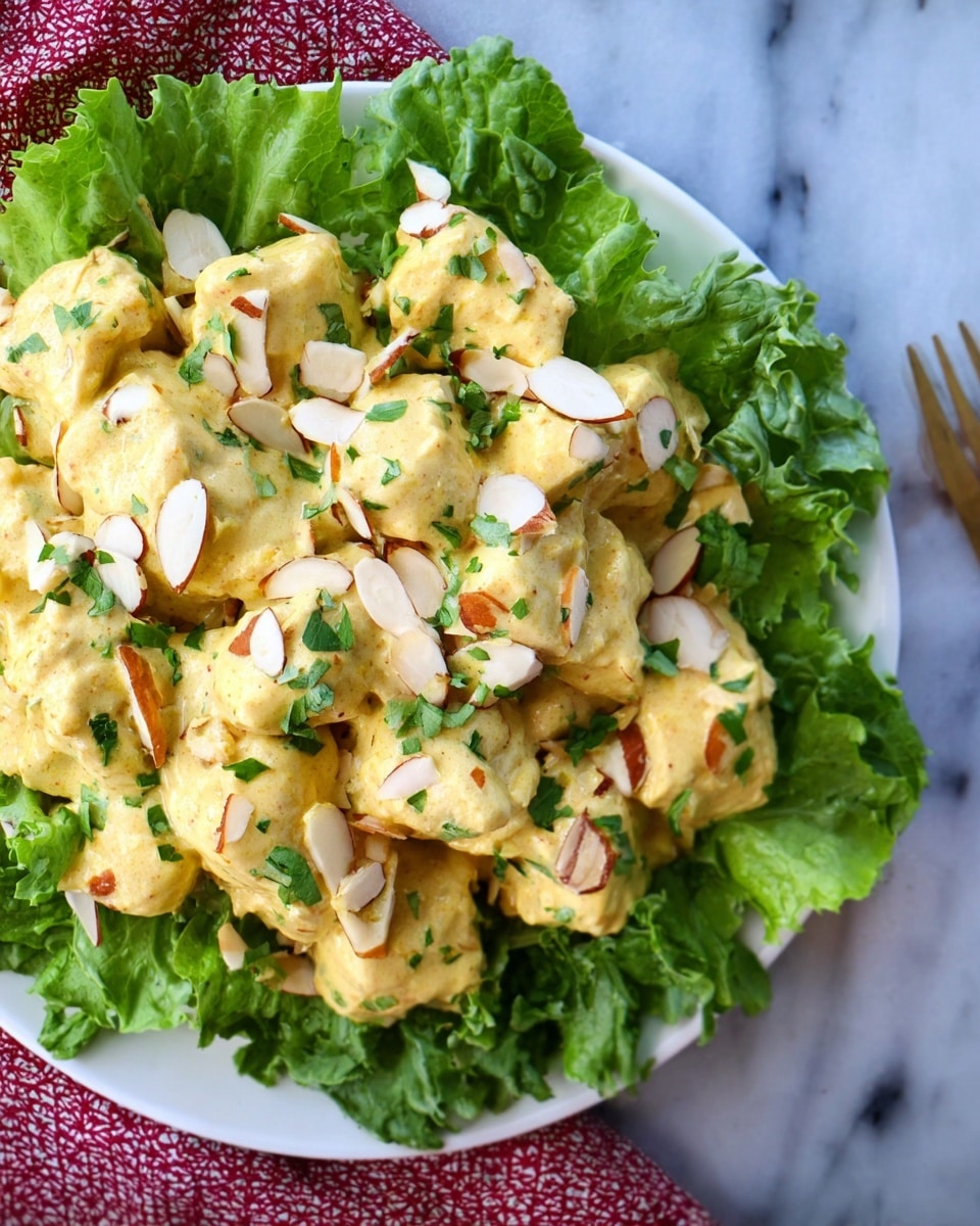 A white bowl holds a fresh bed of green leafy lettuce with ruffled edges, topped with several chunks of yellowish creamy chicken salad mixed with visible spices. The chicken pieces are coated smoothly in the sauce, and thinly sliced almond nuts with a light brown edge are scattered on top. Small green herb leaves garnish the dish, adding contrast to the creamy texture. The bowl is set on a white marbled surface with a hint of a red patterned cloth nearby. Photo taken with an iphone --ar 4:5 --v 7