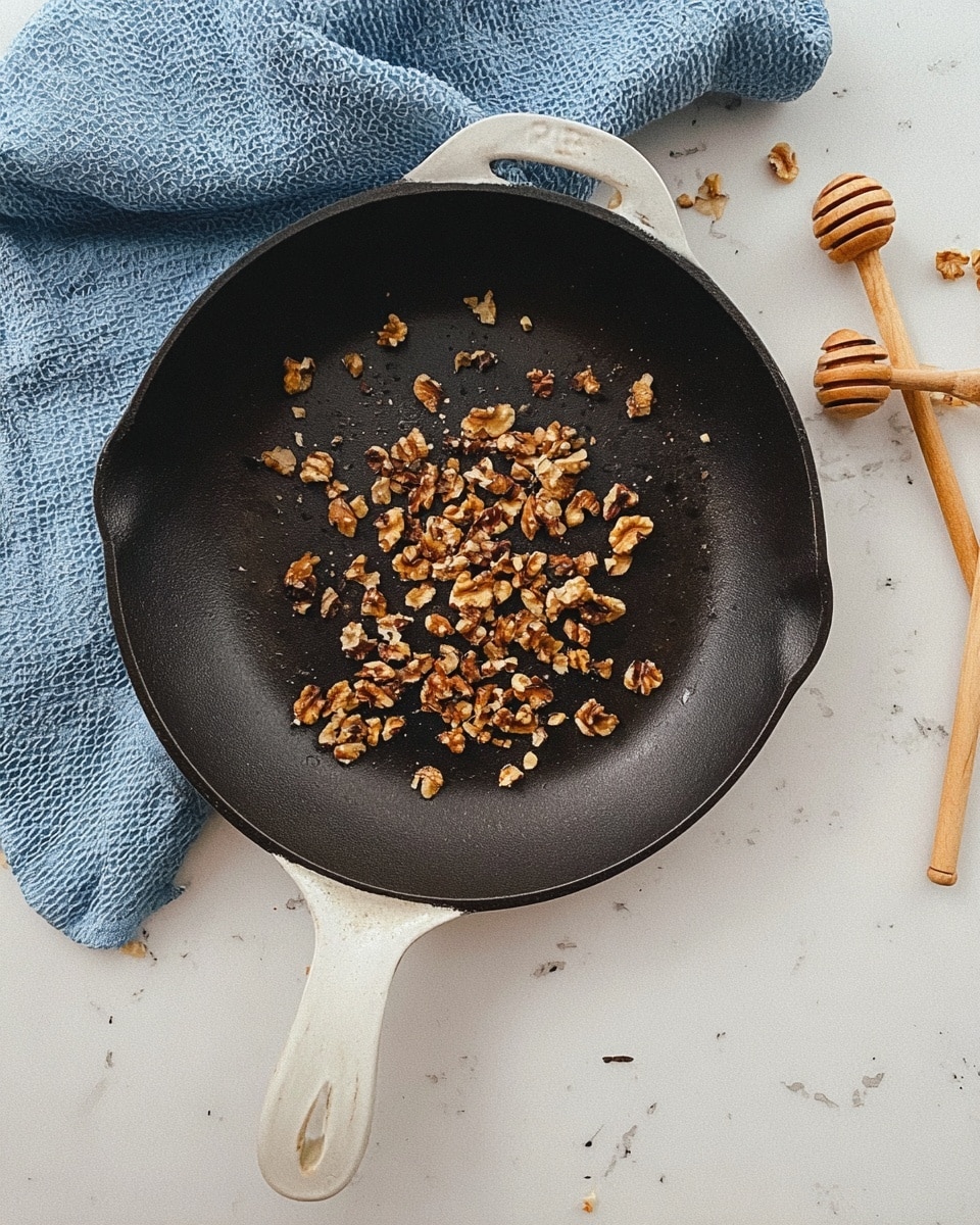 A dark cast iron pan with a white handle holds a scattered pile of small, uneven walnut pieces in the center. The pan sits on a white marbled surface, partly covered by a textured blue cloth to the top left. Near the pan on the right side, there are two light wooden honey dippers laid down side by side. The scene is simple with focus on the walnuts and the pan's worn surface photo taken with an iphone --ar 4:5 --v 7