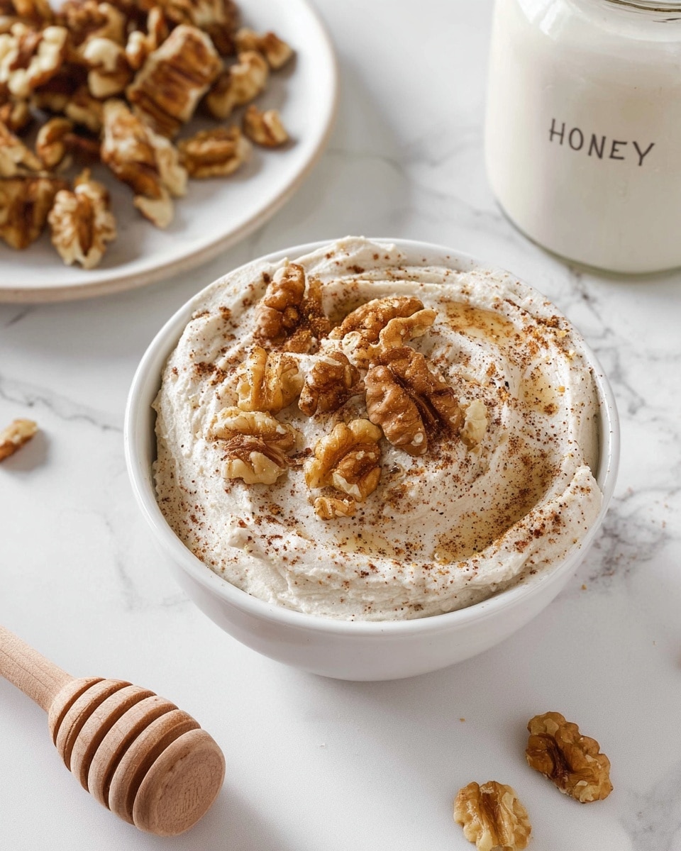 A small white bowl filled with a creamy, swirled mixture that is off-white with tiny brown specks, topped with several whole walnut pieces and a light dusting of brown spice, all sitting on a white marbled surface. Nearby, there is a white plate with more walnut pieces, part of a white jar labeled