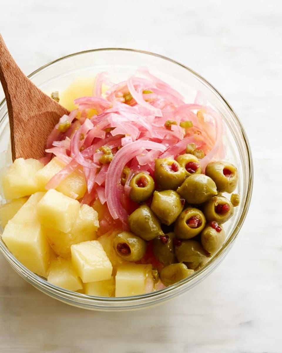 A clear glass bowl is filled with three main layers of ingredients: at the bottom, pale yellow potato cubes form the base with a firm texture; on top of the potatoes are thinly sliced pink pickled onions that are slightly translucent and shiny; to one side, a cluster of green olives stuffed with small red pimentos adds contrast in color and texture. A wooden spoon is partially dipped into the bowl from the left side. The bowl sits on a white marbled surface. Photo taken with an iphone --ar 4:5 --v 7