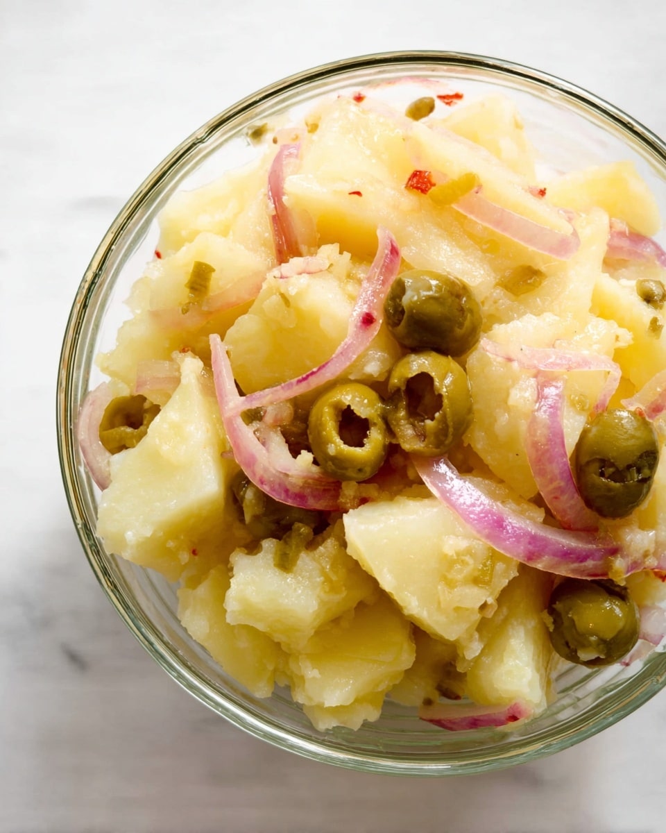 A clear glass bowl filled with thick, chunky pieces of pale yellow potato mixed with thin, long slices of pinkish-red onion and whole green olives with bits of red inside. The potato pieces have a soft and slightly rough texture, while the onions add a light curl and shine with their slightly wet look. The olives are smooth and round, placed evenly throughout the bowl. The bowl sits on a white marbled surface. photo taken with an iphone --ar 4:5 --v 7