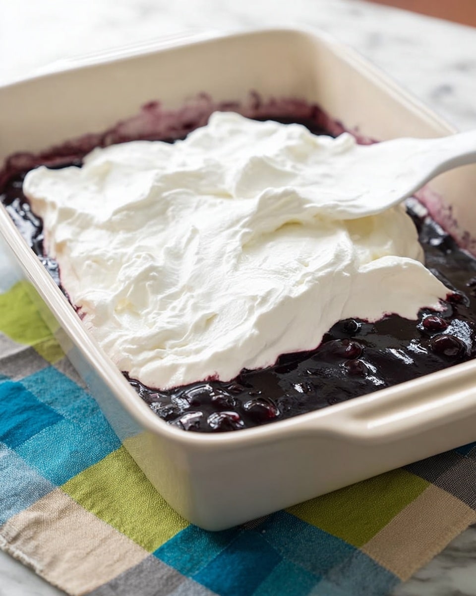 The image shows a white baking dish with two layers of dessert. The bottom layer is a glossy, dark purple-blue mixture with visible small chunks, likely berries or fruit. The top layer is a thick, soft, and fluffy white cream spread unevenly over the fruit layer. A white spatula rests on top, partly submerged in the cream, with texture marks from spreading. The dish is placed on a colorful cloth with green, blue, beige, and gray squares over a white marbled surface. Photo taken with an iphone --ar 4:5 --v 7