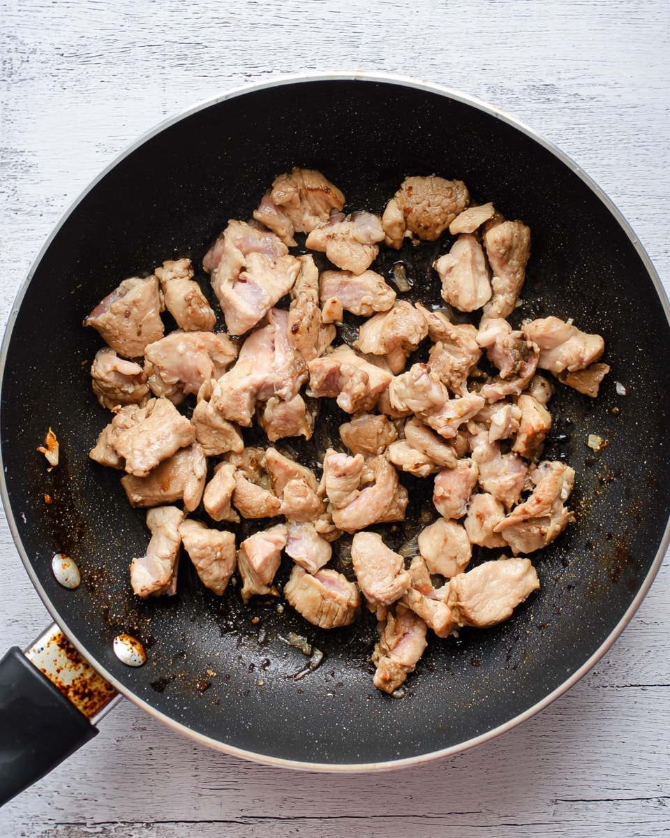 The image shows a black frying pan with many small, uneven pieces of cooked light brown chicken scattered inside. The chicken pieces have a browned texture on top and some pinkish-white inside. The pan has a black handle on the left side with some rust near where the handle meets the pan. The pan sits on a white marbled surface with faint gray wood-like markings. photo taken with an iphone --ar 4:5 --v 7